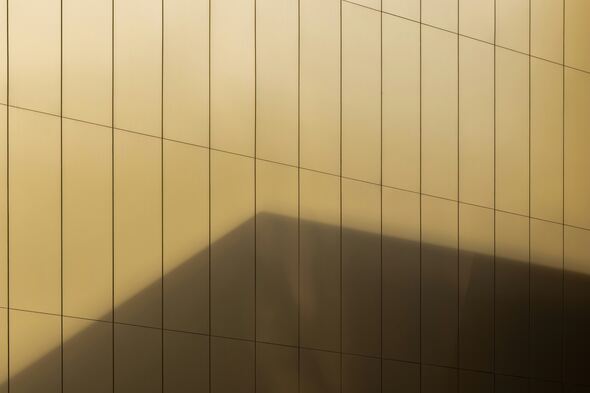 A minimalist architectural facade composed of vertical and horizontal panel lines on a golden surface, with a large shadow cast diagonally across the wall, creating geometric contrast.