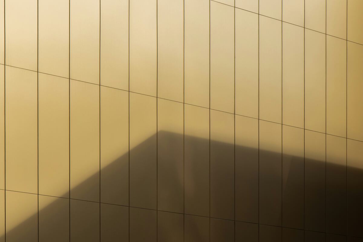 A minimalist architectural facade composed of vertical and horizontal panel lines on a golden surface, with a large shadow cast diagonally across the wall, creating geometric contrast.