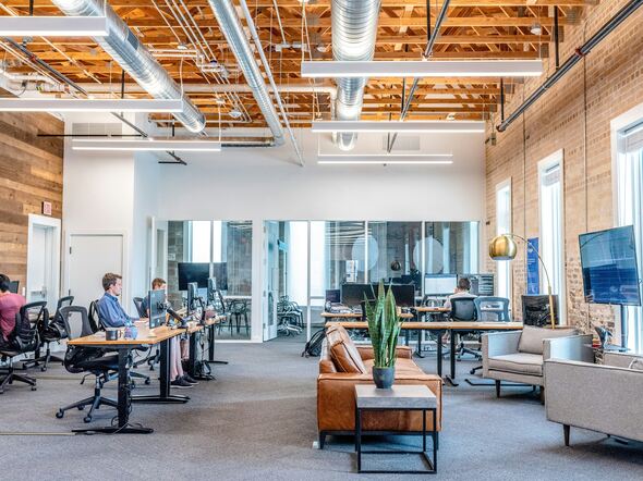 Bright open-plan coworking space with exposed brick walls, modern furniture, and professionals working at shared desks in a tech startup office.