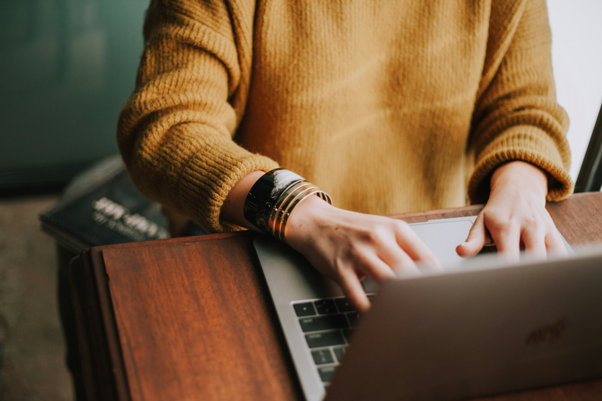 Close-up photo of a person's hands typing on a laptop keyboard. The person is wearing a mustard yellow sweater and multiple bracelets on their wrist, which rests on a wooden desk. There are blurred books to the side, indicating a cozy and studious environment.