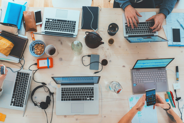 A bird's-eye view of a busy worktable. Several individuals appear to be engaged in various tasks, predominantly using laptops. The table is littered with numerous items indicative of a collaborative work environment: there are at least four laptops, notebooks, a pair of headphones, a teapot with cups, a bowl of snacks, and mobile phones. Chargers and a portable hard drive suggest that the work involves digital content. The setting is casual and contemporary, likely a co-working space or a creati