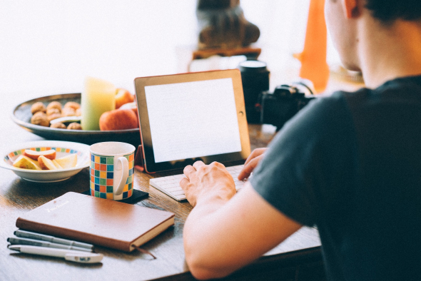 a photo take from the back of a man typing on ipad with a keyboard on what may seem a dining table with coffee mug a notebook plates of fruit and a blurred view of a camera far behind