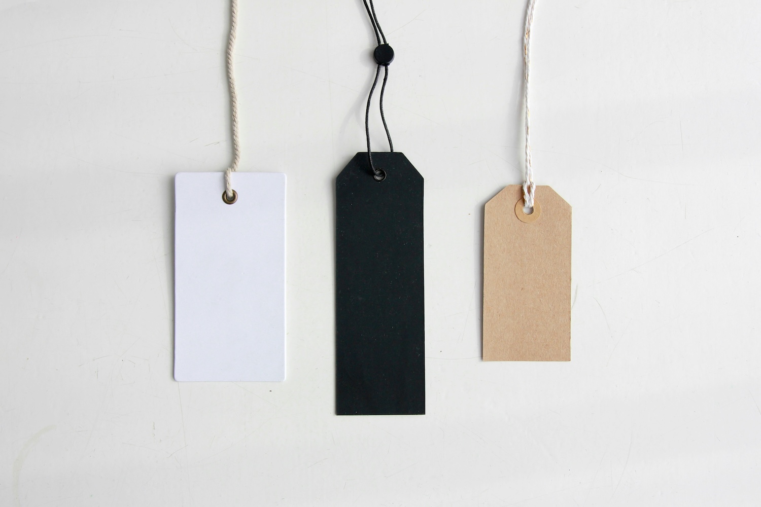 Three blank product tags in white, black, and kraft paper hanging against a white background.