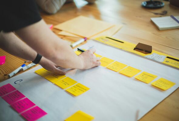 Person organizing yellow sticky notes on a large white sheet during a brainstorming or planning session, with markers and documents on a wooden table.