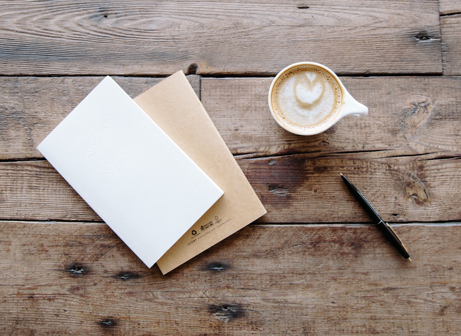 an overhead shot of a notebook to the left and a cup of coffee and a pen all laid out on top of a rustic wooden table