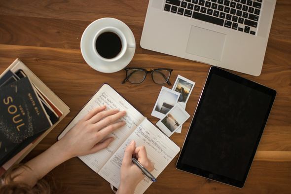 Overhead view of a person writing in a journal with a cup of coffee, a pair of glasses, a stack of books, a laptop, an iPad, and a few instant photos on a wooden table.