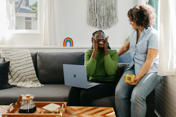 A person is sitting on a gray sofa with a laptop on their lap, laughing while covering their face with hands, as another person stands beside them, hand on their shoulder, both enjoying a lighthearted moment. There is a coffee table with a carafe and cups in the foreground, and a woven wall hanging and rainbow decoration in the background.