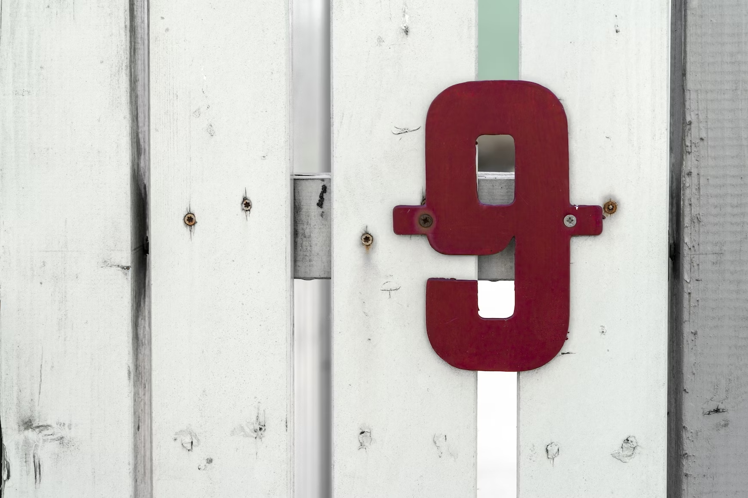 Red number 9 mounted on a weathered white wooden fence with visible screw heads and a vertical turquoise strip.