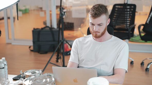 A young man with short hair and a beard, wearing a white t-shirt, sits focused at a table working on a silver MacBook laptop. The background shows an office space with chairs, equipment cases, and a tripod, with food containers and drinks on the table in the foreground.